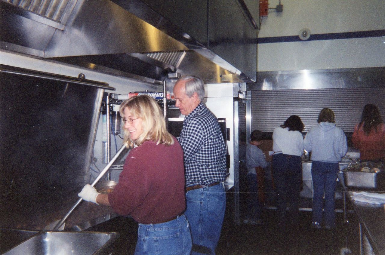 People in a commercial kitchen preparing food. Woman stirs pot, man watches. Others work at a counter.