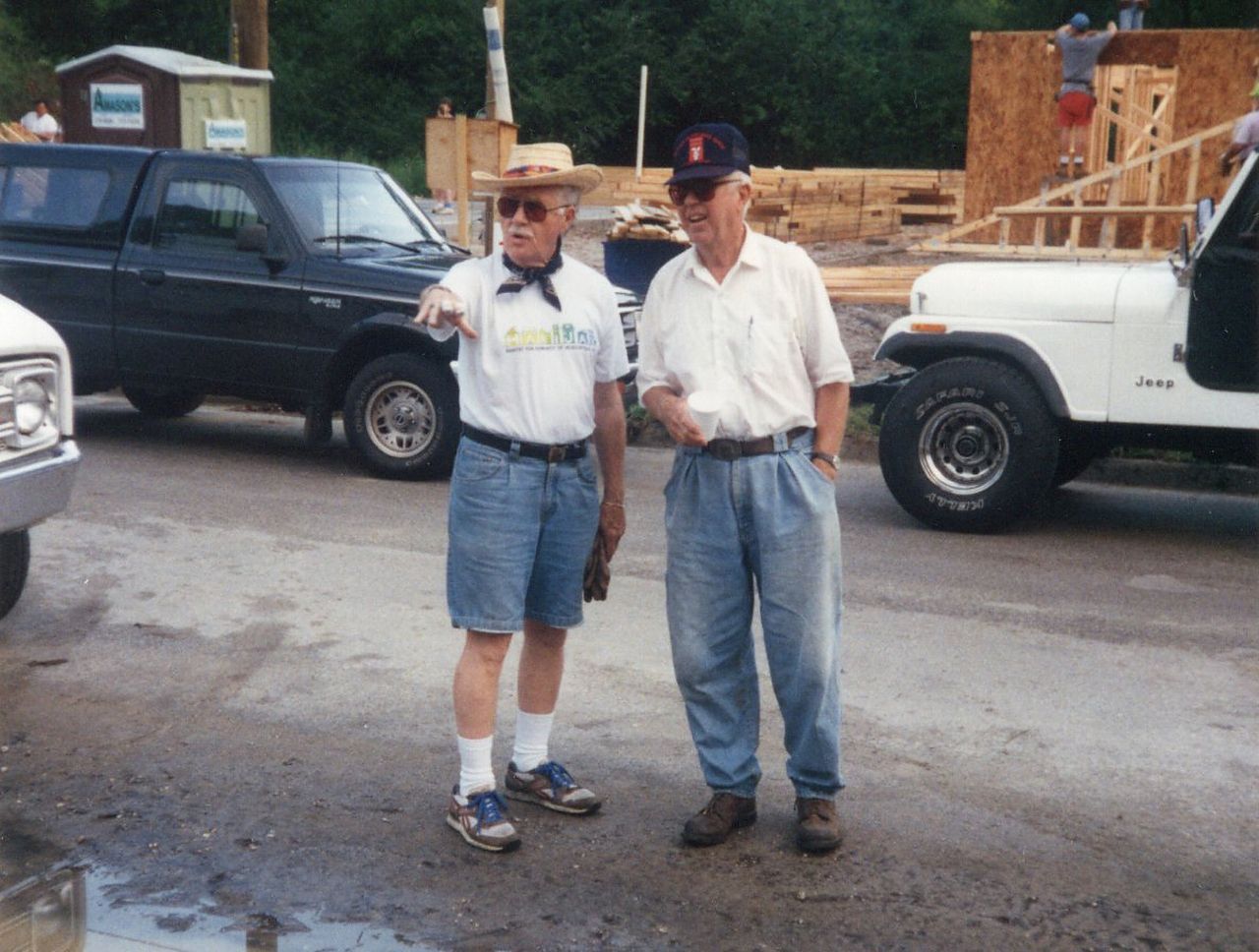 Two men standing outside near vehicles and a construction site. One points, the other has hands in pockets.