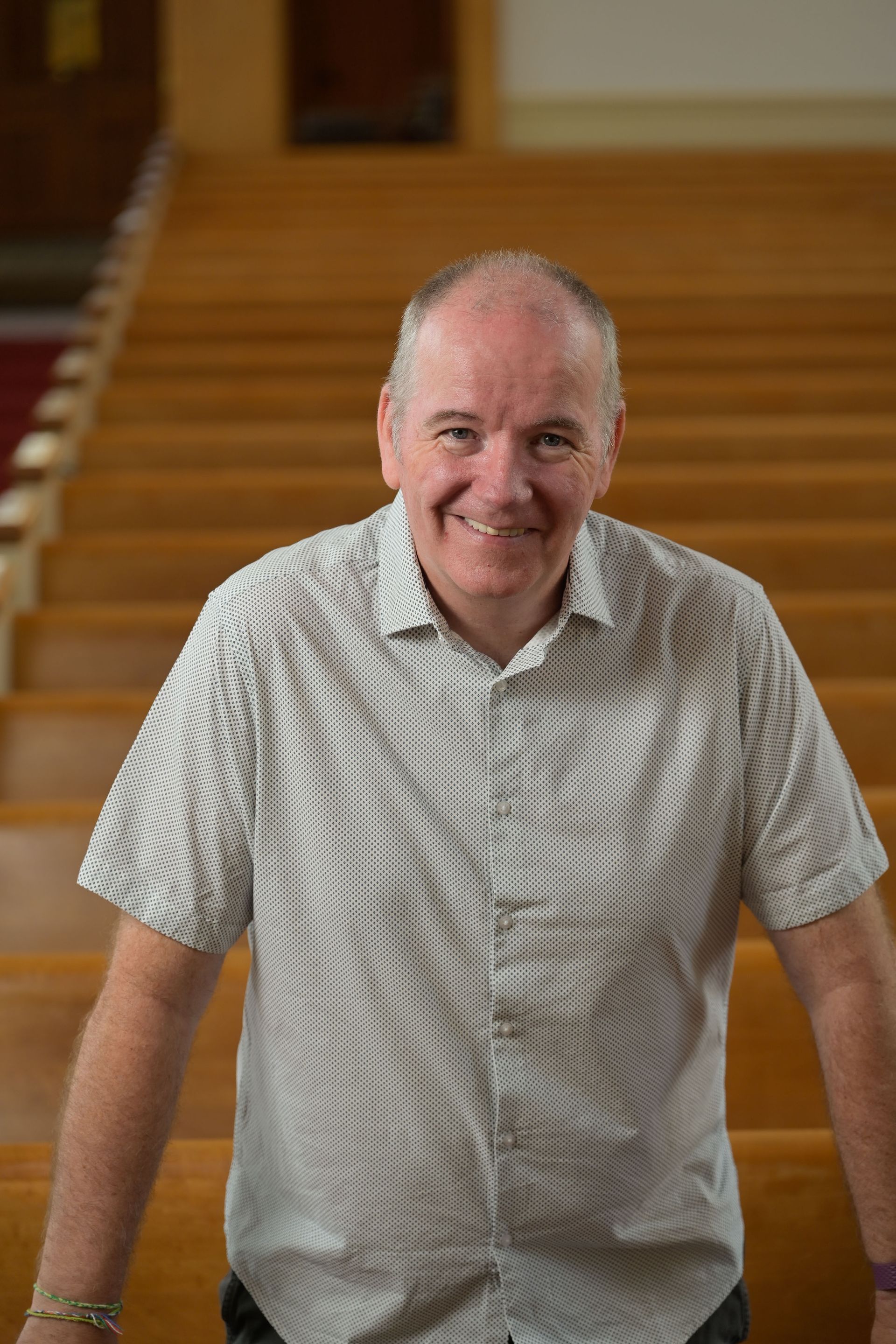 Man smiling, standing in a church with rows of wooden pews. He wears a patterned shirt.