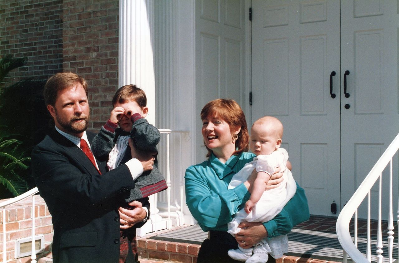 Family stands in front of a white church: father in suit, mother holding baby, older child crying.