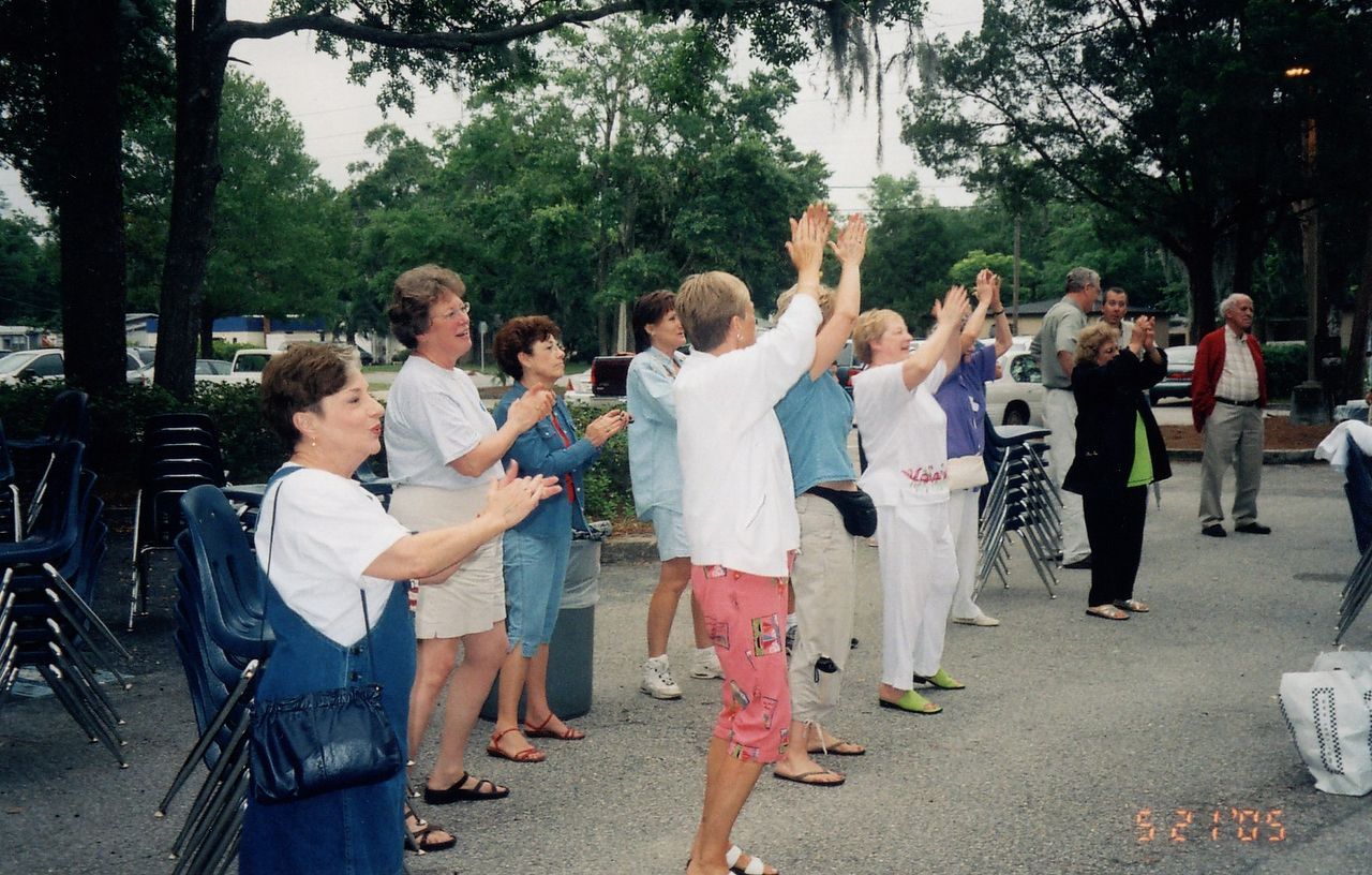 People clapping and raising arms outdoors; trees and parked cars in background.