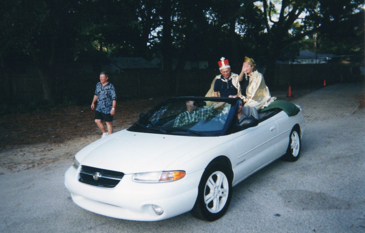 Three people in a white convertible car, two wearing festive hats.  A person walks alongside. Roadside setting.