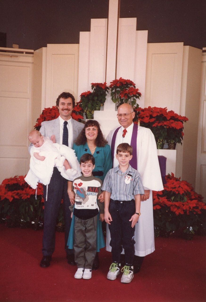 Family and a clergyman pose for a photo in a church. A baby is held by a man wearing a suit.