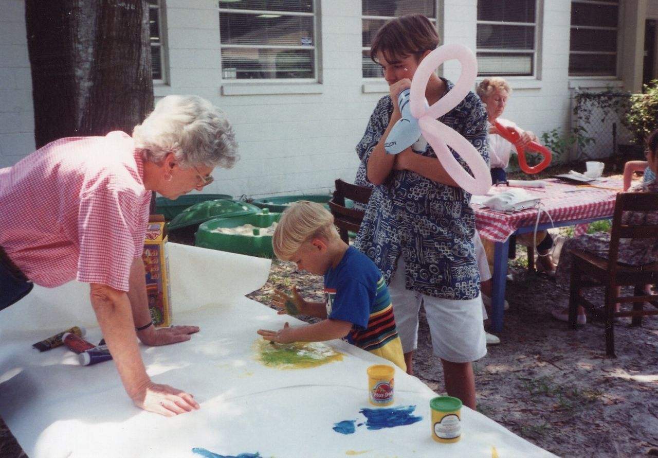 Woman and child painting at an outdoor table; another woman holds a balloon.