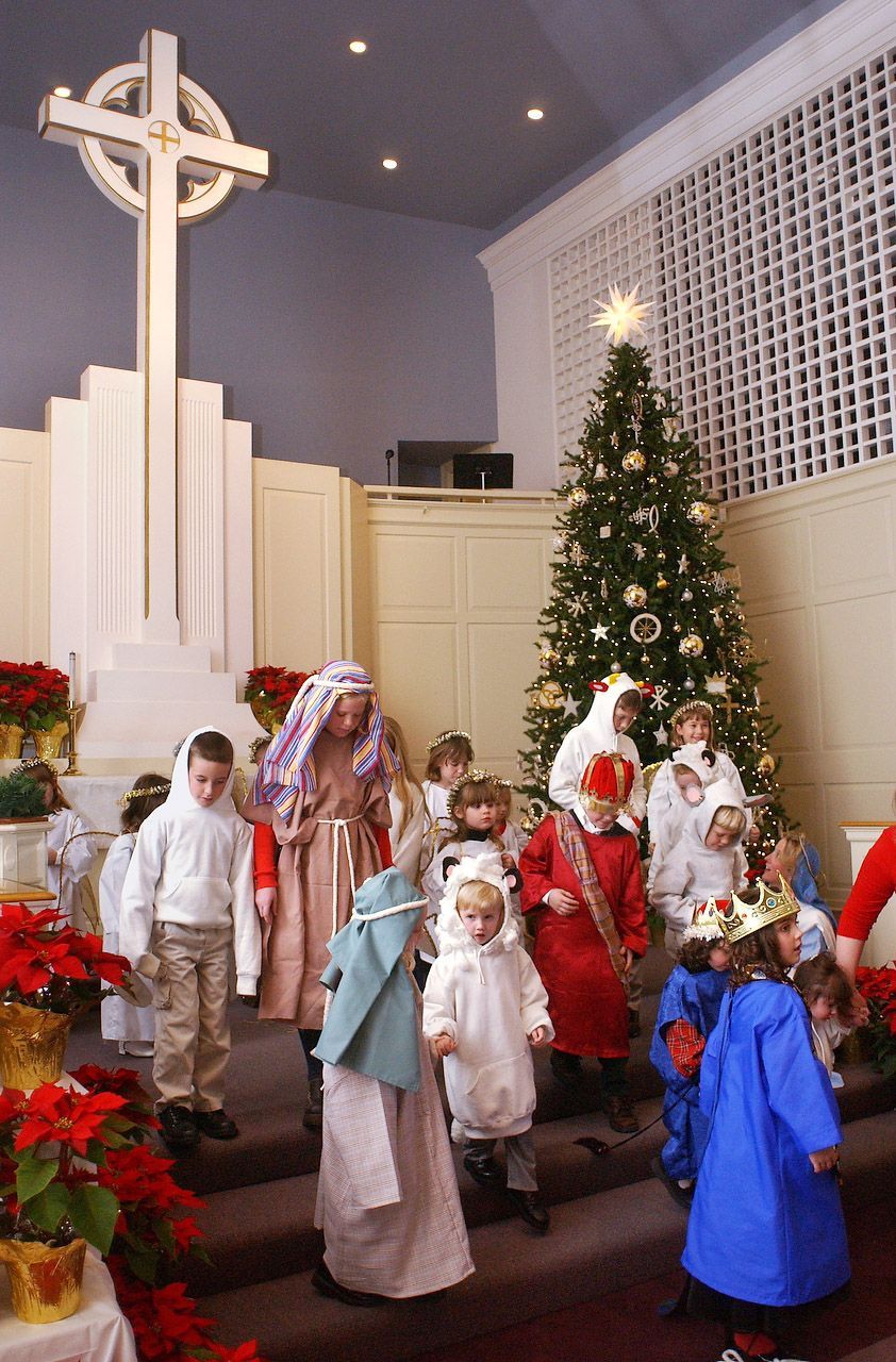 Children in costumes walk down steps near a Christmas tree and a cross in a church.