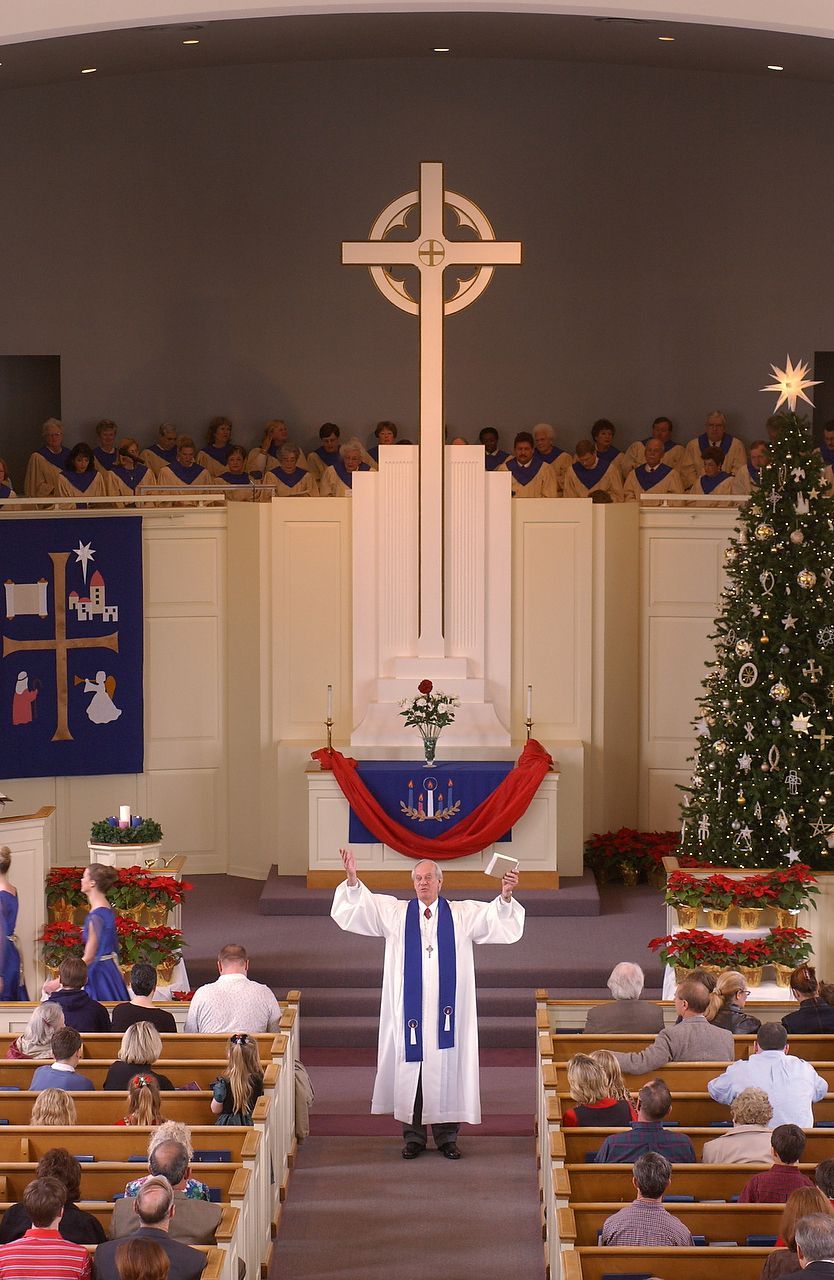 Pastor in white robe raises arms in church, with cross, choir, and Christmas tree.