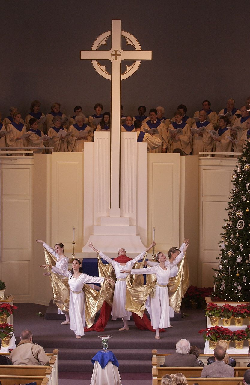 Dancers in white robes and gold capes perform on stage below a large cross in a church. A choir is in the background.