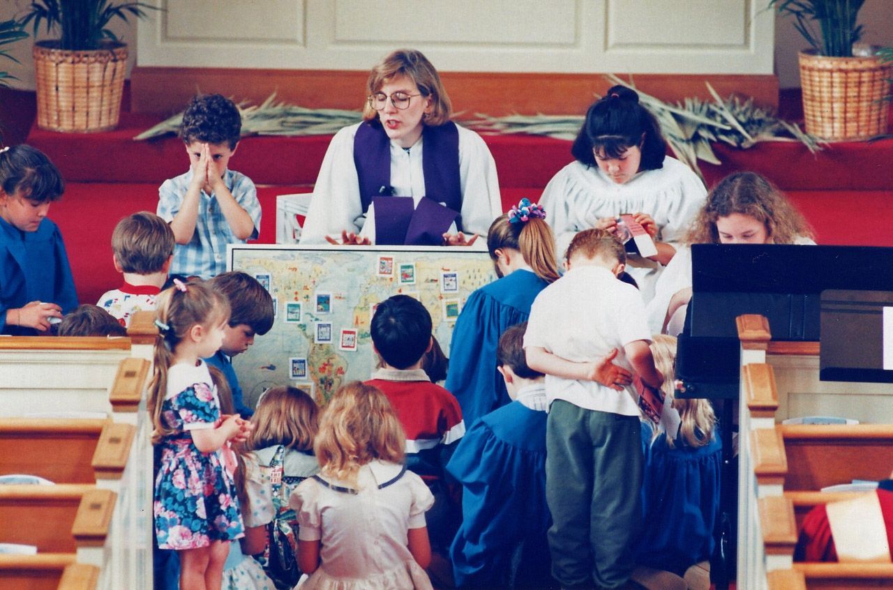 Children and adults gather around a map, a woman in a purple vestment speaks in a church.