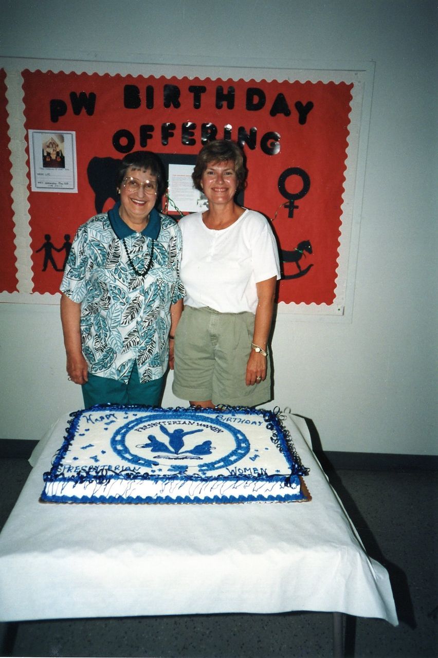 Two women stand behind a cake with a blue logo. A bulletin board with text is visible.