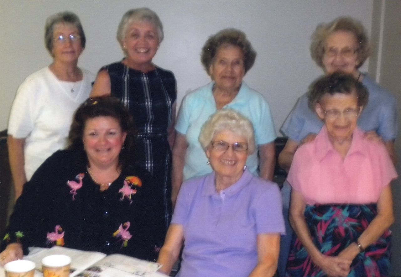 Group of women smiling, gathered around a table; some seated, others standing against a wall.