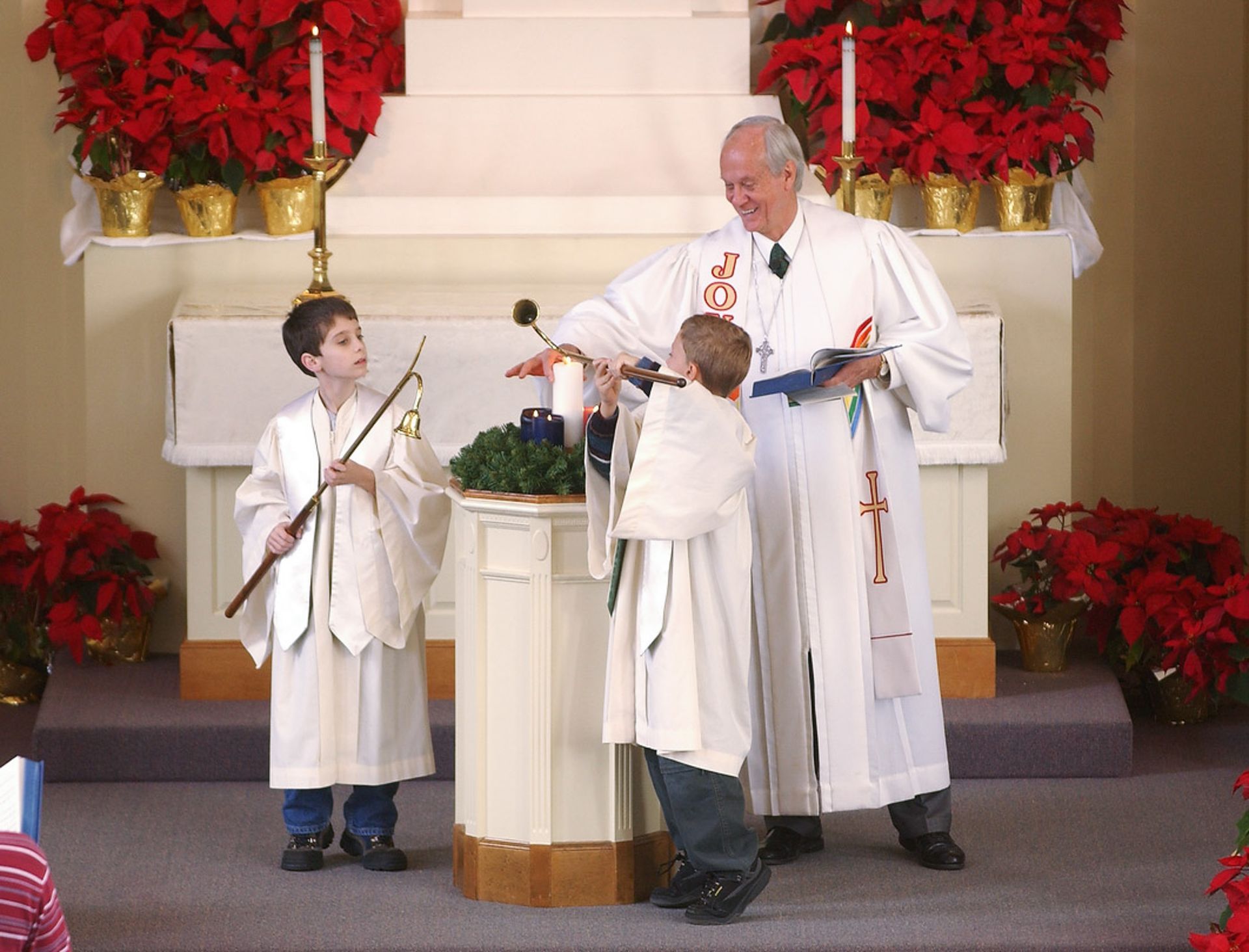Pastor and two children lighting Advent wreath candles at altar. Church interior, poinsettias.