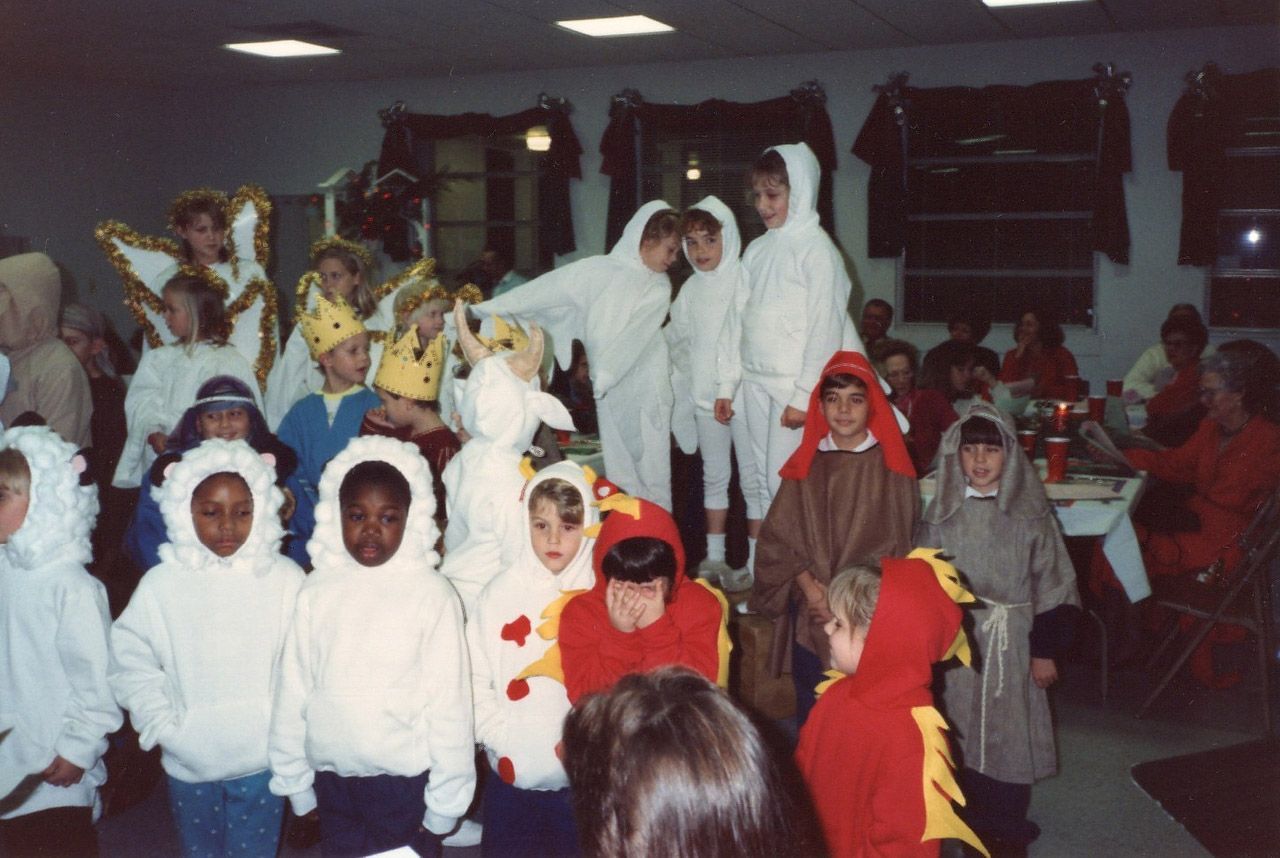 Children in costumes, likely for a play. White and red hooded outfits, some with animal designs. Indoors, festive setting.