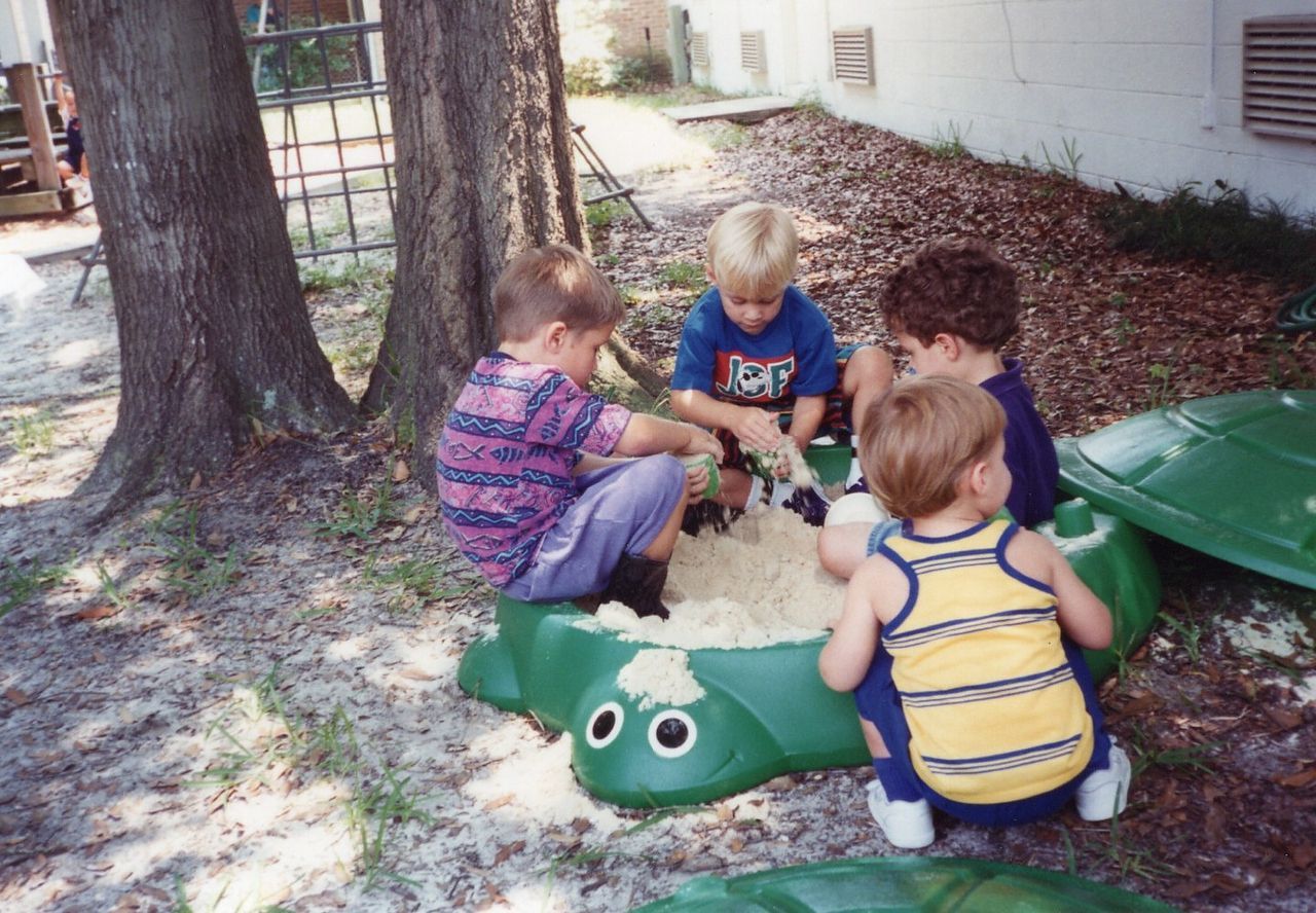 Four children playing in a green turtle-shaped sandbox filled with sand.