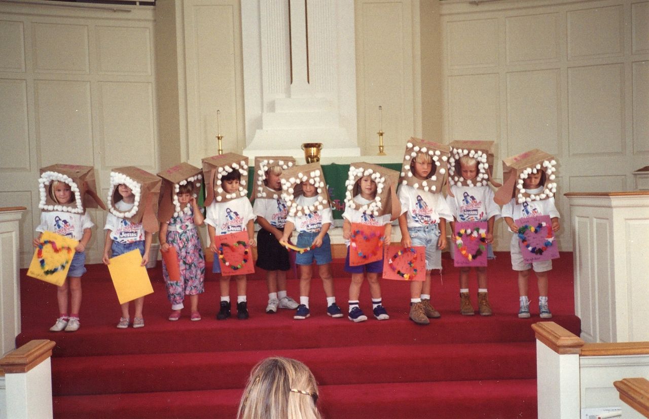 Children wearing bread slice costumes stand on a stage in a church. Each holds a decorated sign.