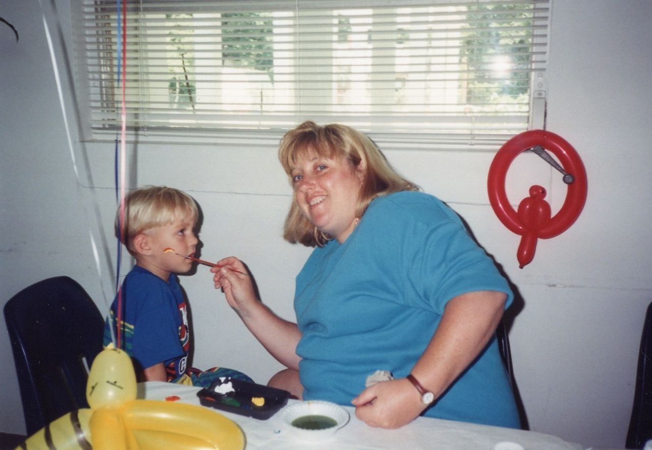 Woman applies face paint to a young child indoors at a party. A yellow balloon is nearby.