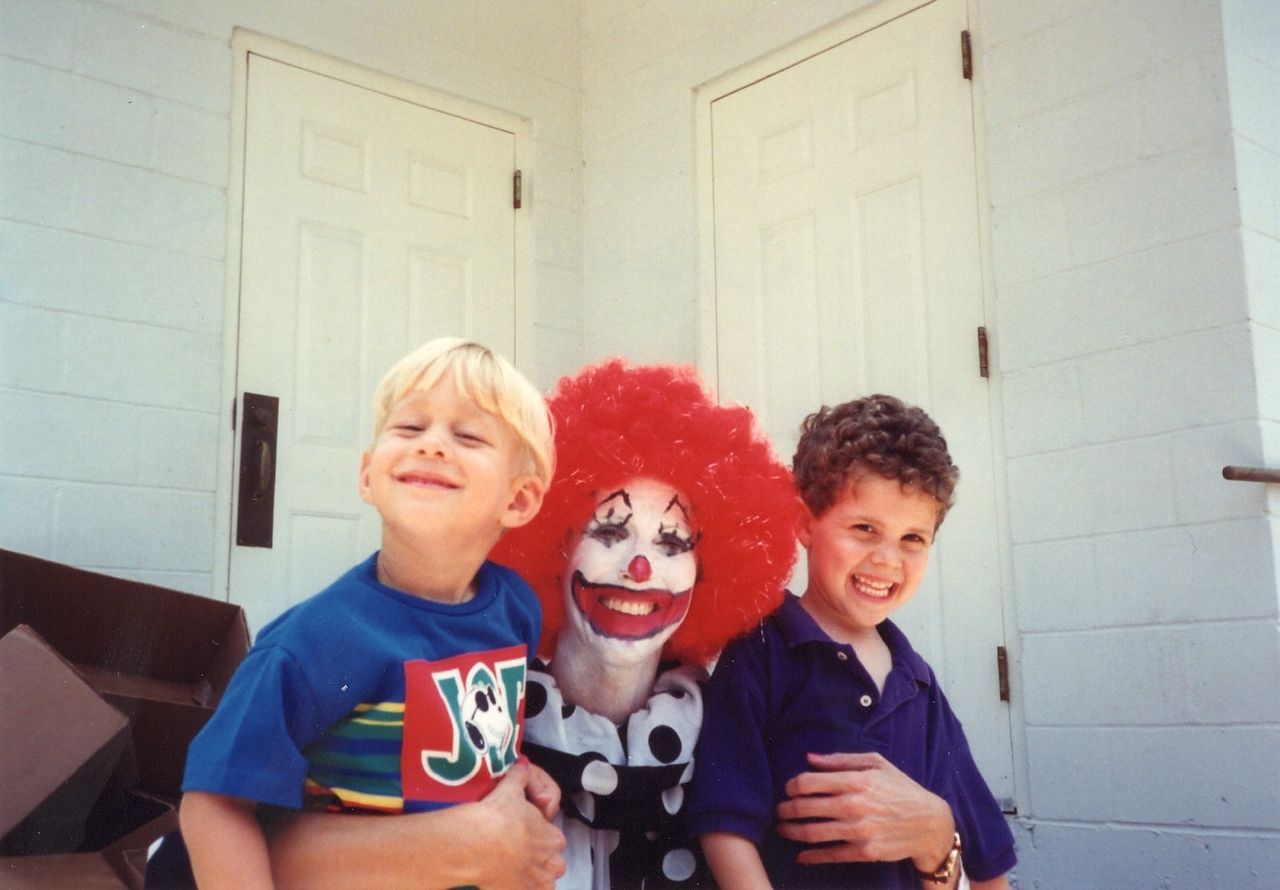 Clown with red hair and painted face hugs two smiling children in front of white doors.