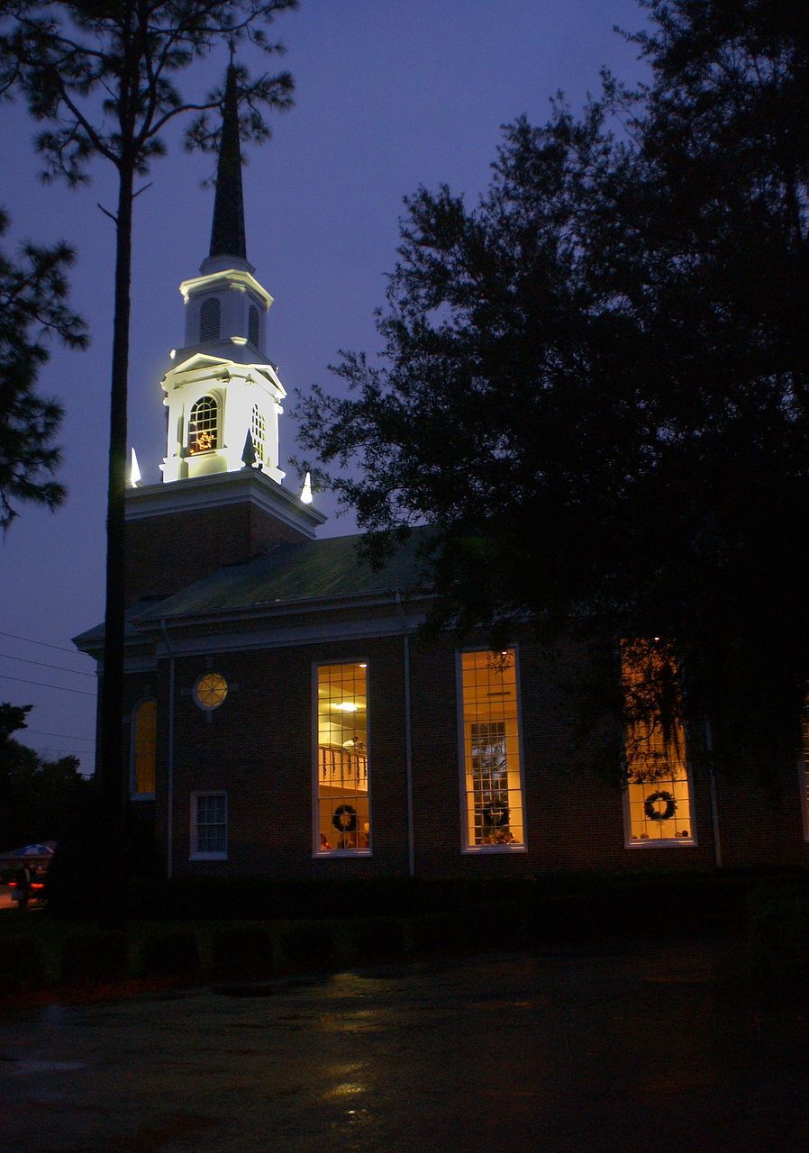 Brick church with steeple illuminated at night, wreaths in lit windows, trees frame the structure.