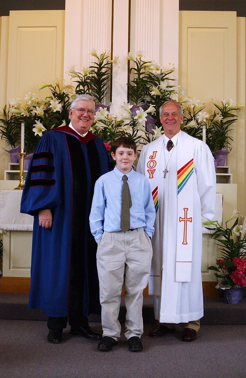 Three people in front of altar with flowers. Man in blue robe and another in white robe with a child.