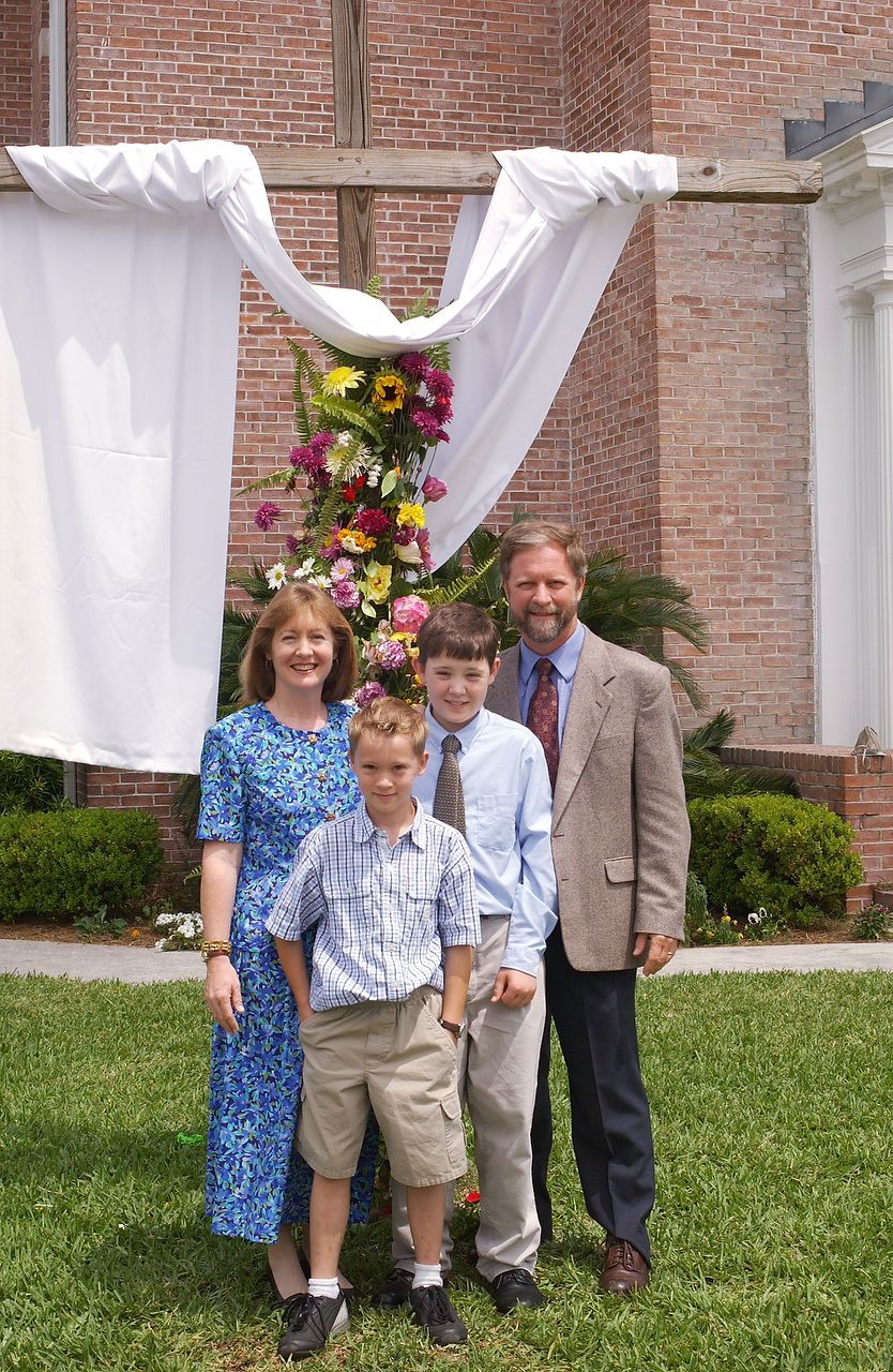 Family posing in front of floral cross and brick building. Woman in blue dress, man in brown jacket, and two boys.