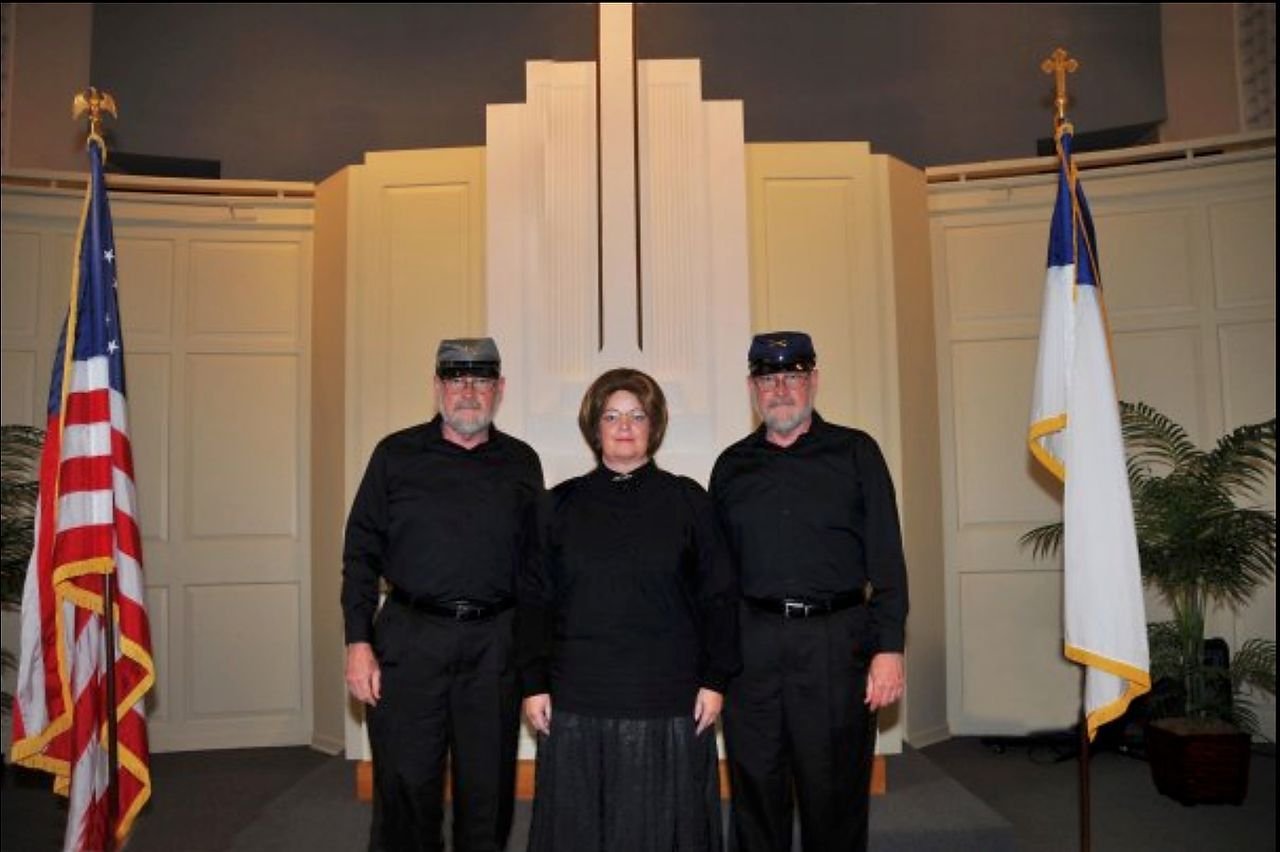 Three people in dark attire stand before a church altar; two wear caps and hold flags.