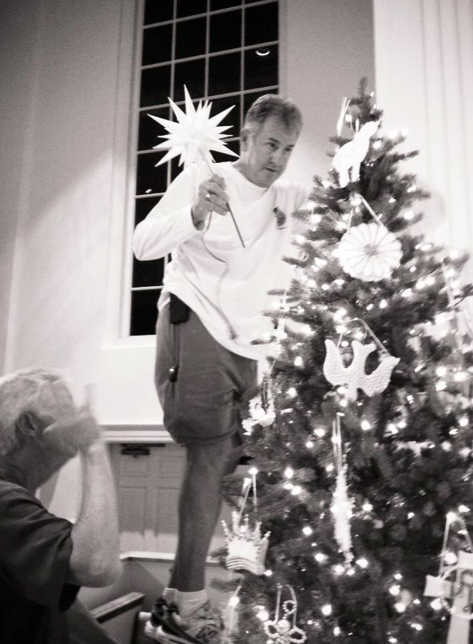Man on a box placing a star on top of a Christmas tree. Another man watches nearby.