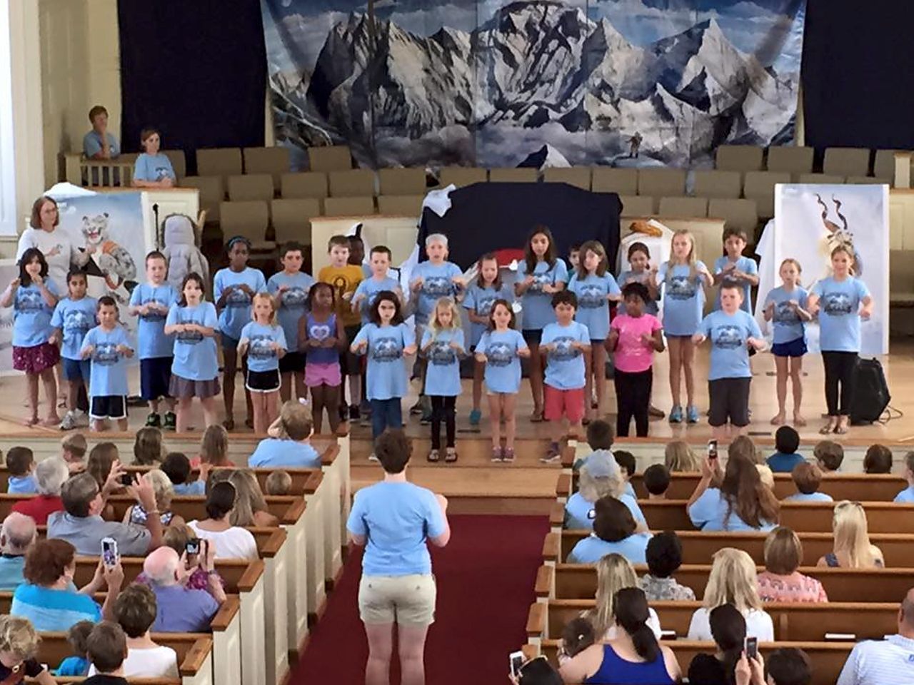 Children's choir on stage, holding items. Audience in pews. Blue, white, and red backdrop of mountains.