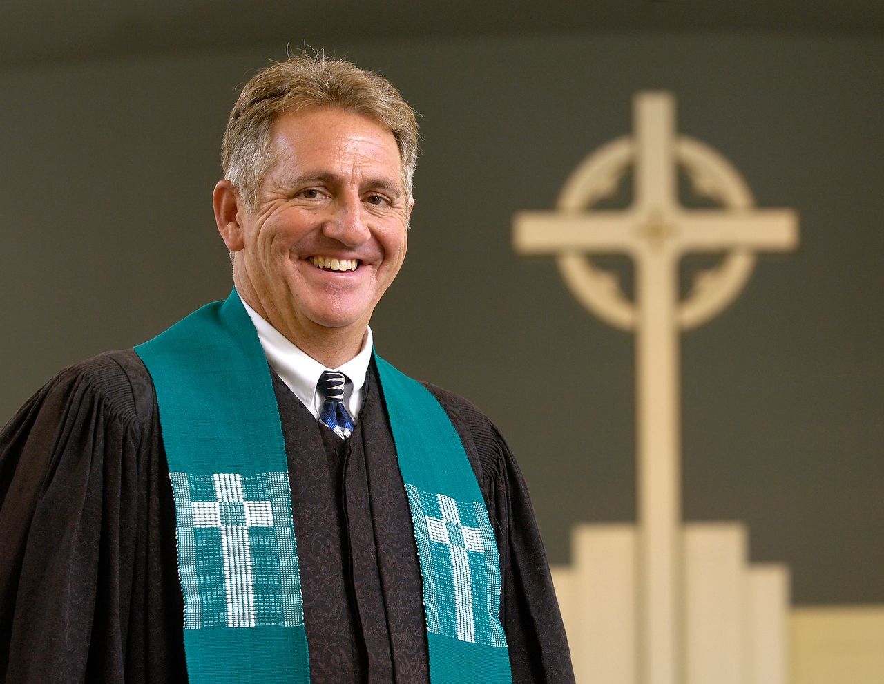 Man wearing a robe and teal stole, smiling in front of a cross.