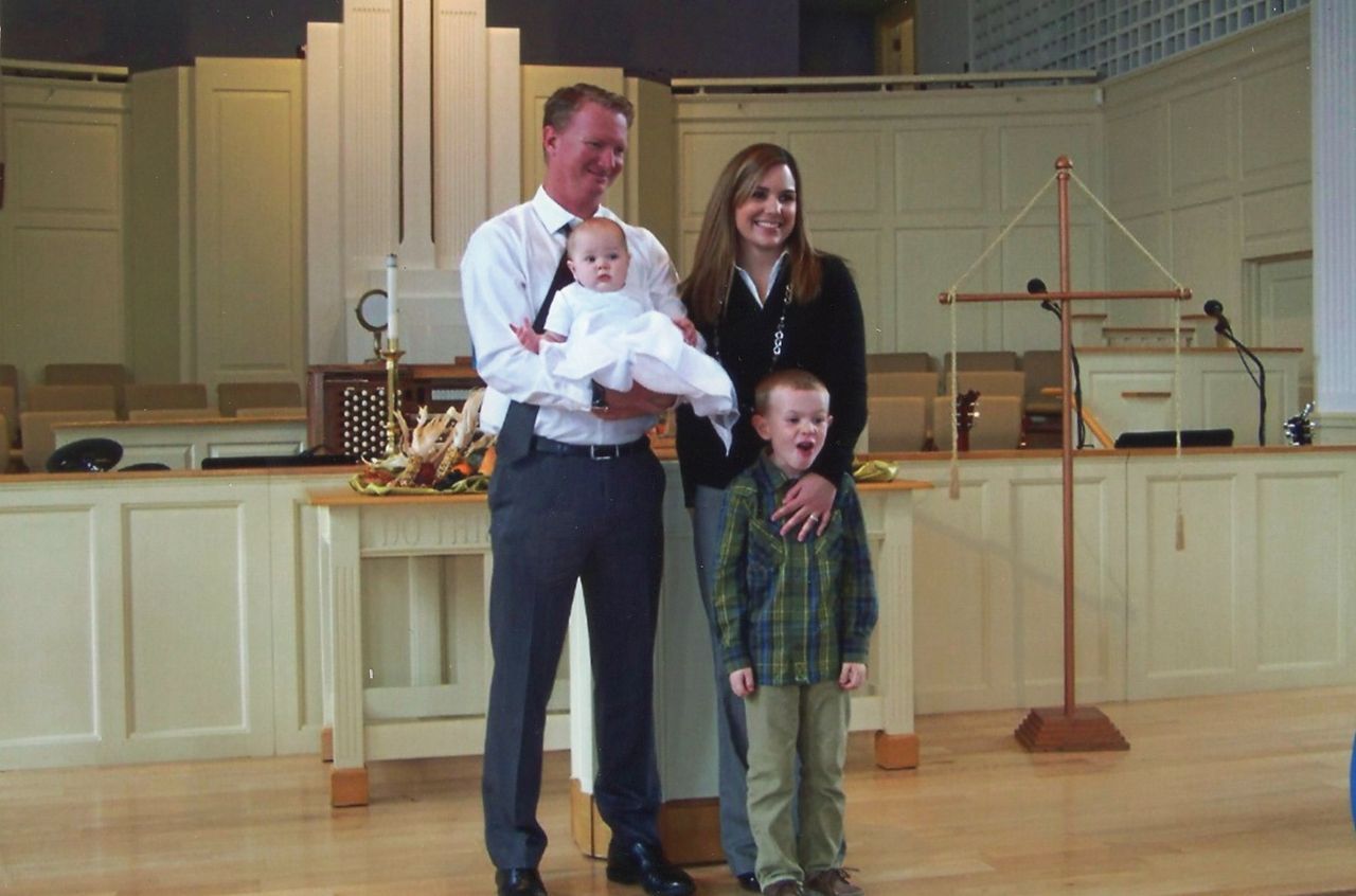 Family of four poses in a church. Man holds baby, woman stands next to older child, all smiling.
