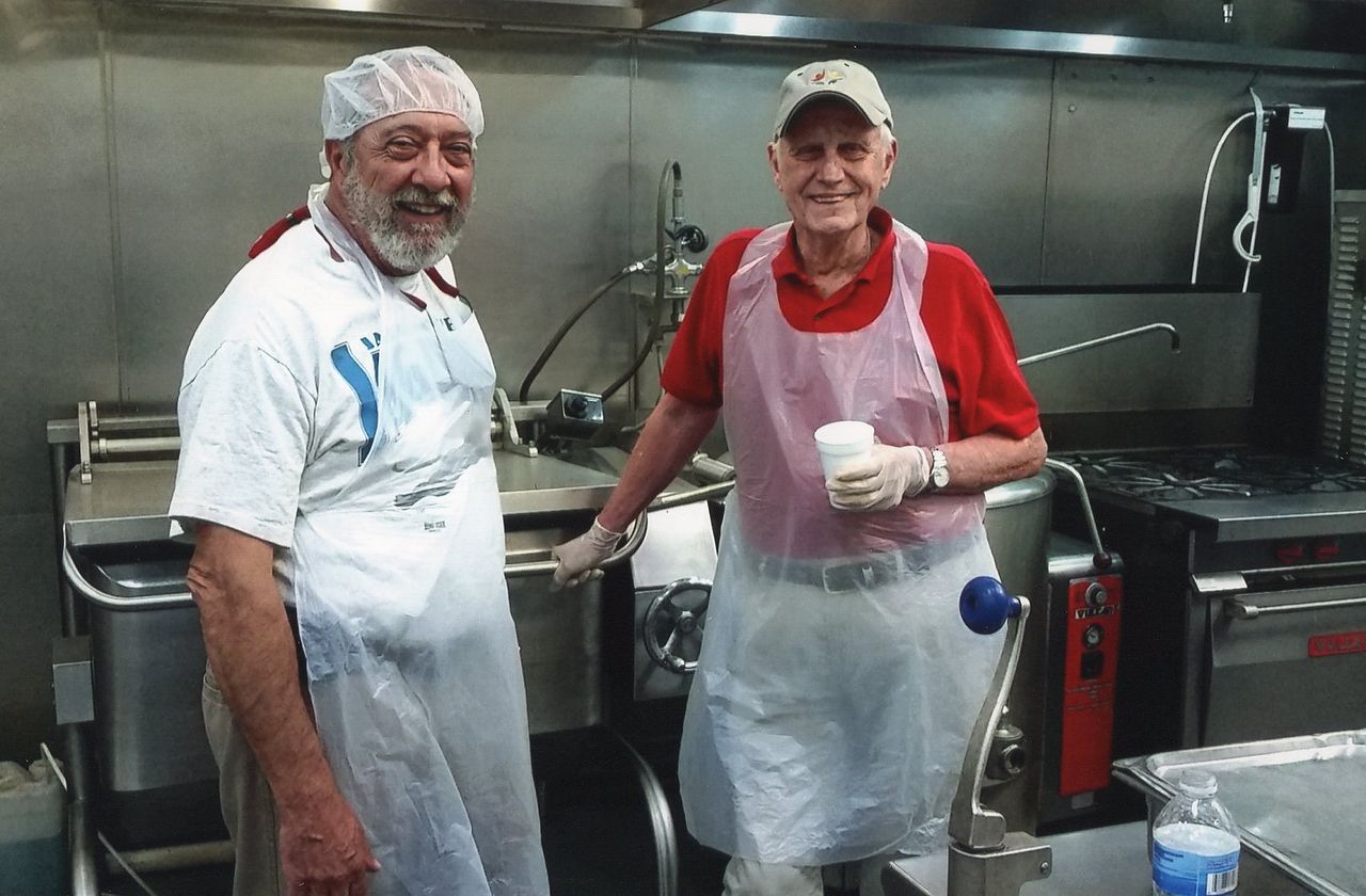 Two men in a commercial kitchen stand by cooking equipment, wearing aprons and hairnets/hats.