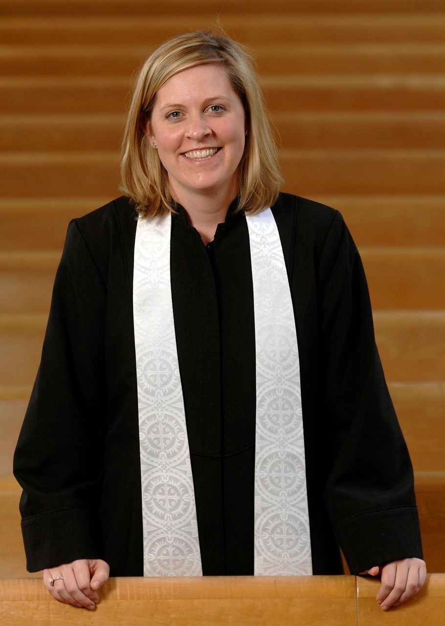 Woman in black robe and white stole, smiling, in front of wooden church pews.