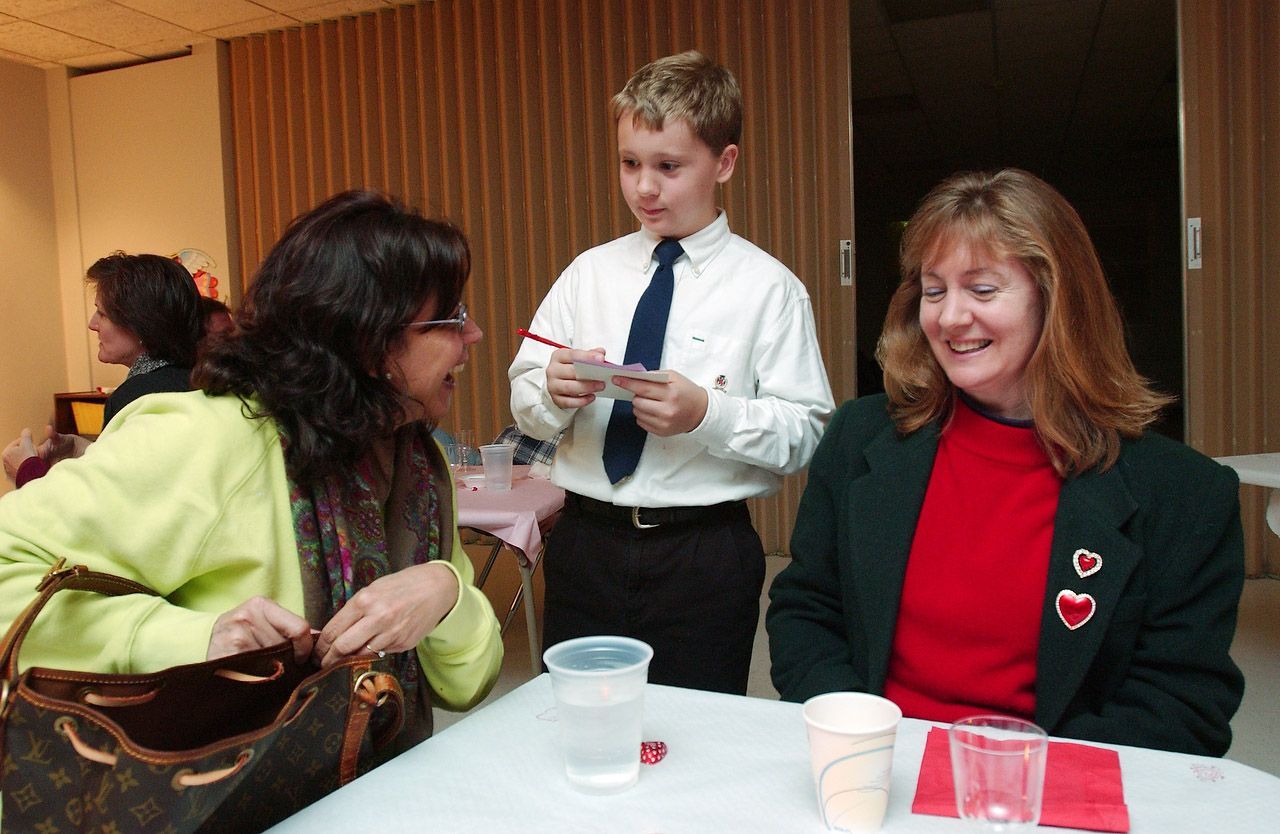 Boy with pen stands between two women at a table. They are looking at him and smiling, possibly playing a game.