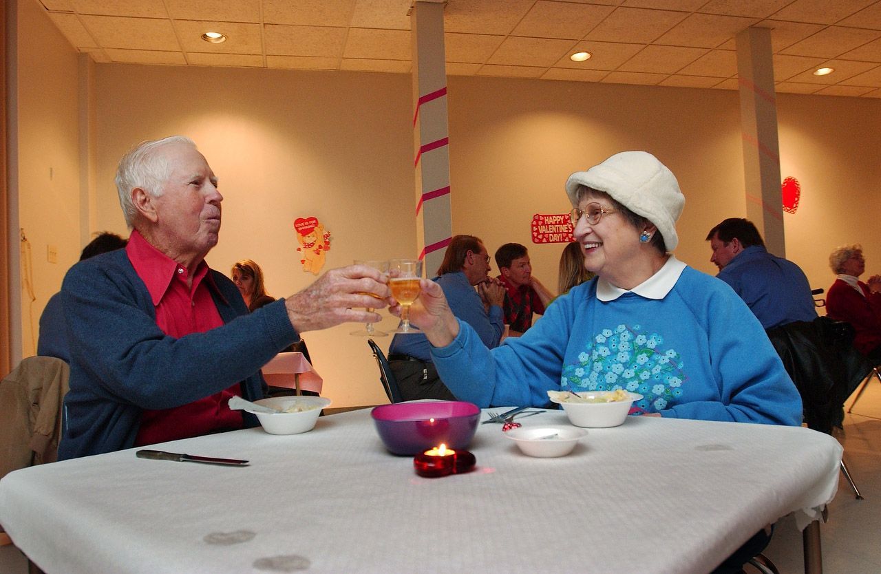 Couple toasting drinks at a table in a room with others. Man in red shirt, woman in blue sweater and hat.