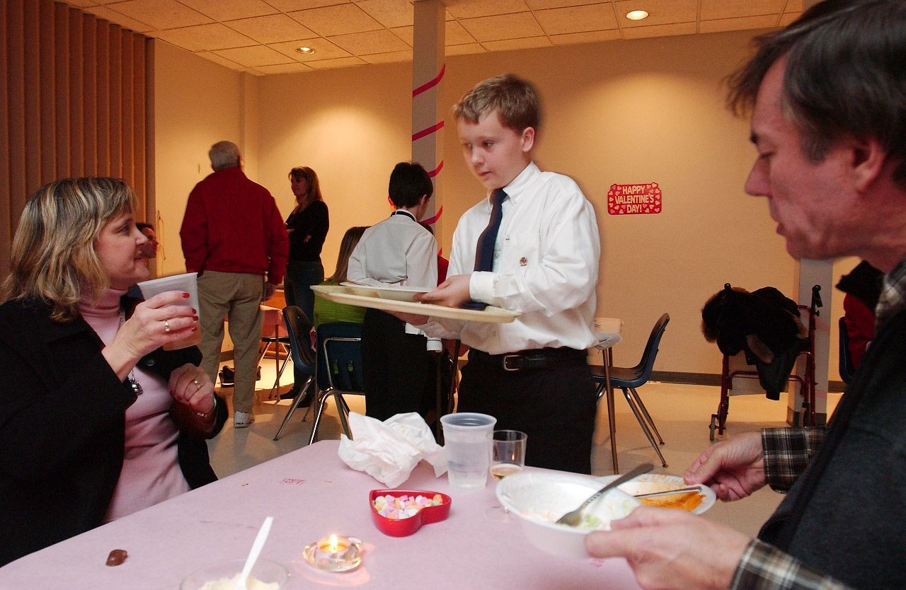Boy in white shirt serves food to people at a table in a gathering room. Others stand in the background.