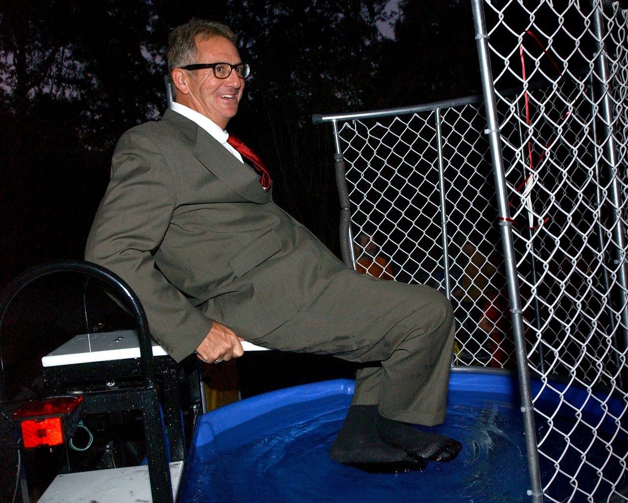 Man in a suit smiles, sits on a dunk tank platform, about to be dunked.
