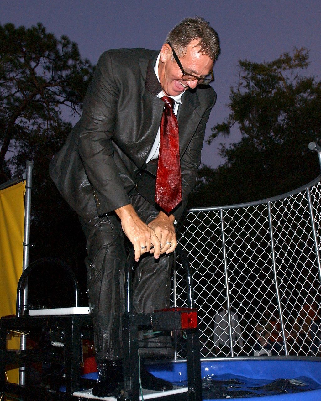Man in a wet suit and tie, smiling, standing in a dunk tank, about to be submerged.