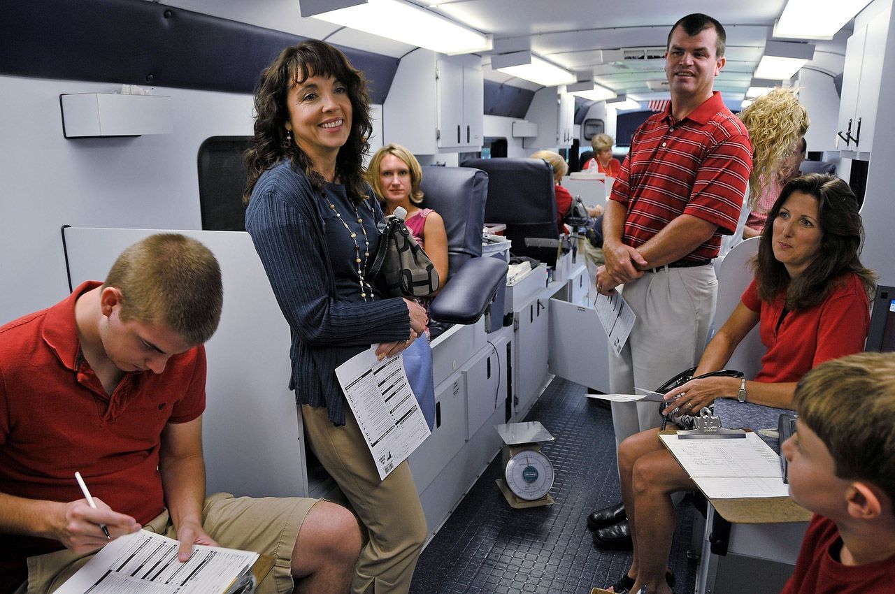 People inside a vehicle; some in red shirts fill out paperwork, others smile at the camera. Interior setting with equipment.