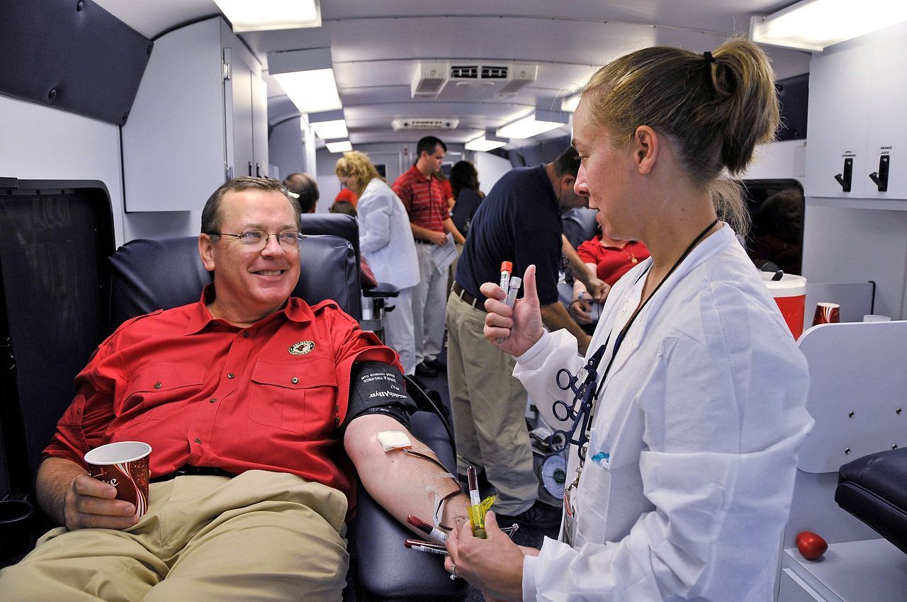 Man in red shirt donating blood in a mobile unit; nurse monitors the process.