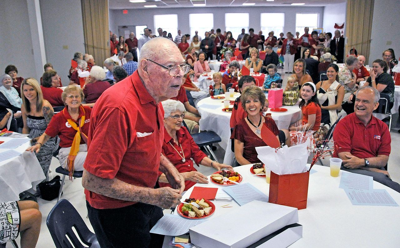 Man in red shirt speaks at a red-themed event with many people, round tables, and decorations in a large room.