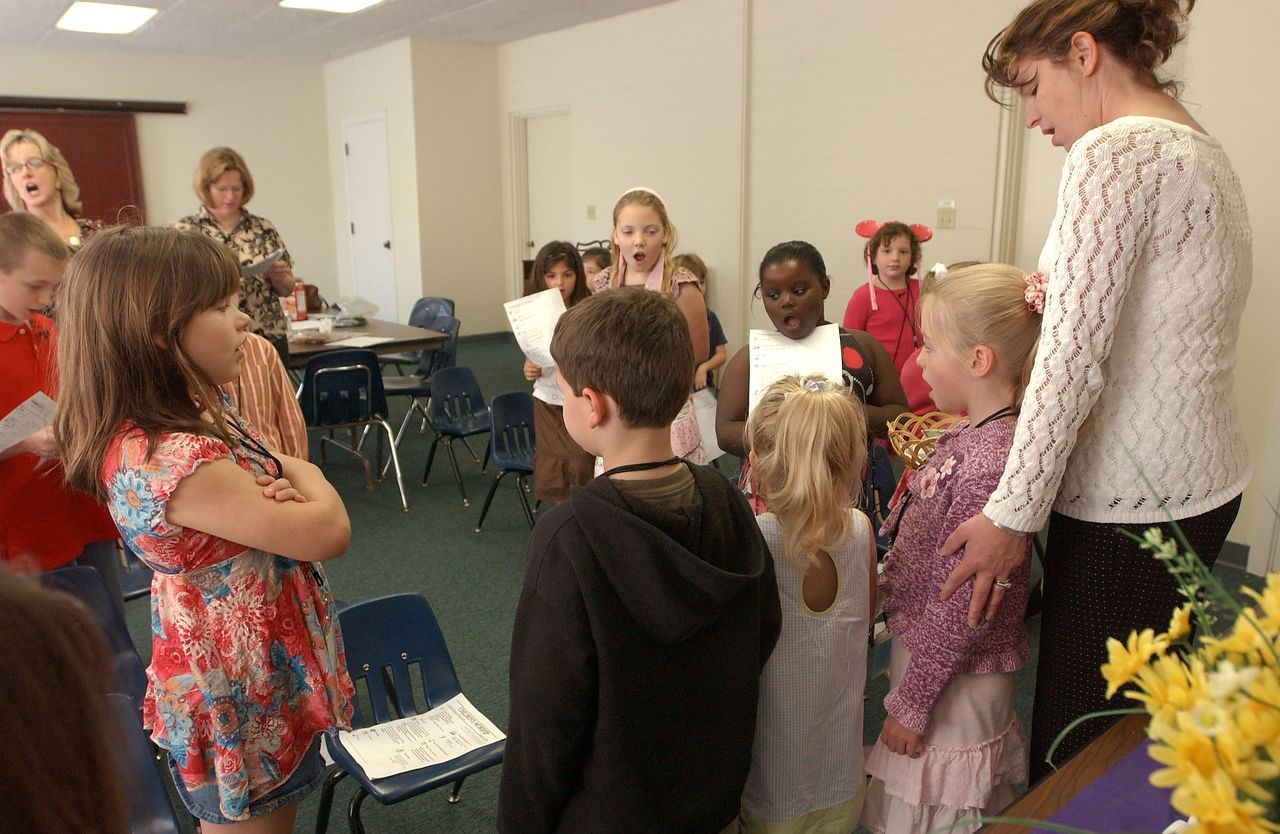 Children in a room with adults, some holding papers, likely rehearsing or performing a song.