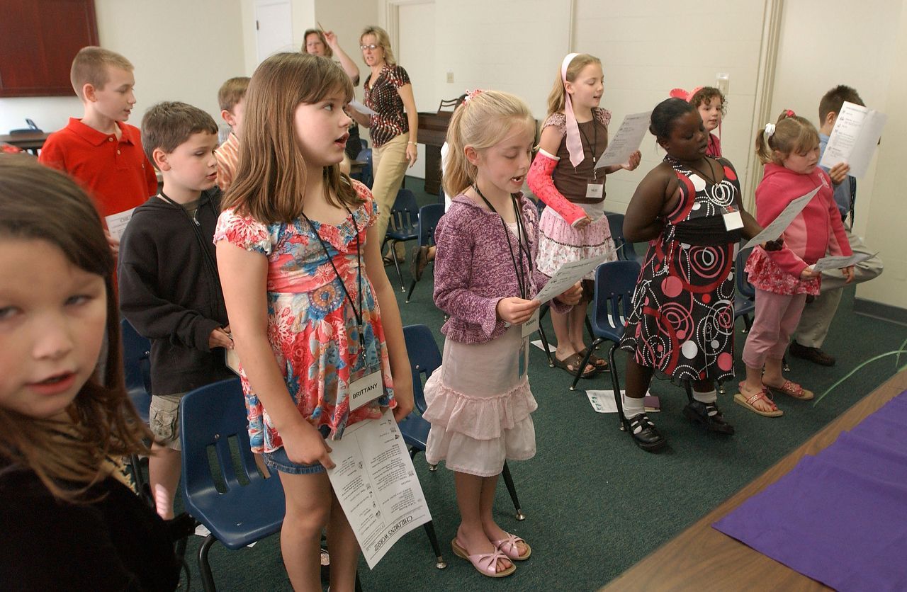 Children singing in a room, holding song sheets, some smiling.