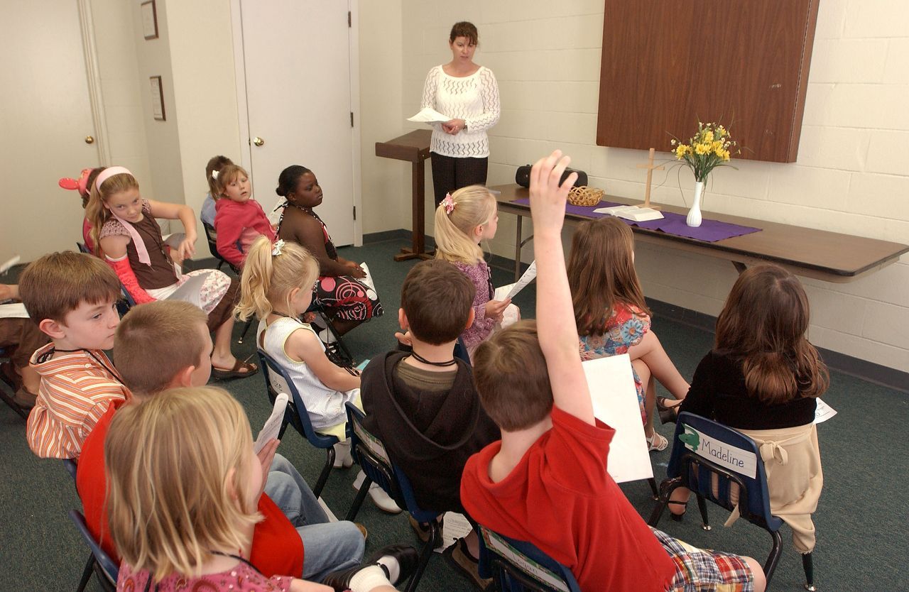 Children in a classroom listen to a woman reading from papers. Some children raise hands.