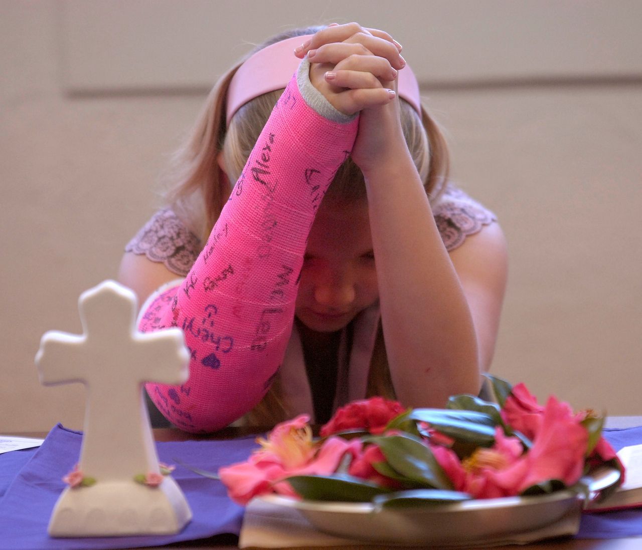 Girl with pink cast praying at a table with a cross and flowers.