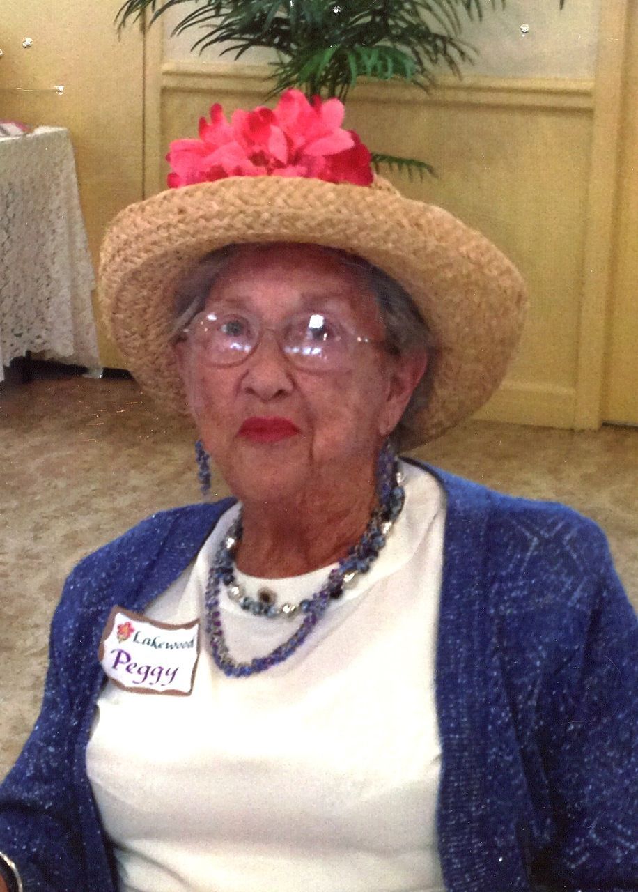 Woman wearing a straw hat with a pink flower, glasses, and a blue cardigan, at an event.