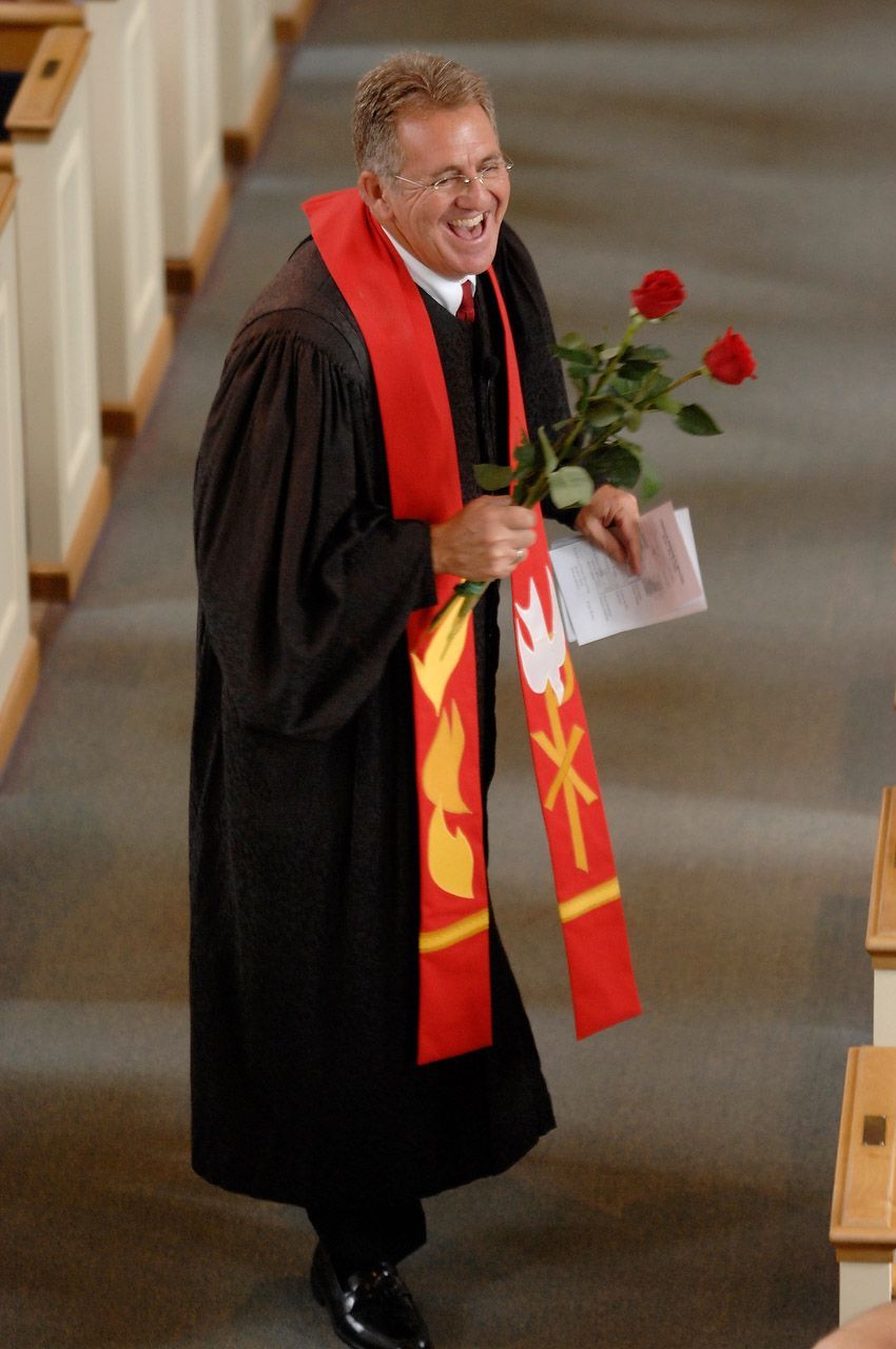 Man in black robe with red stole, holding roses, smiling in a church.