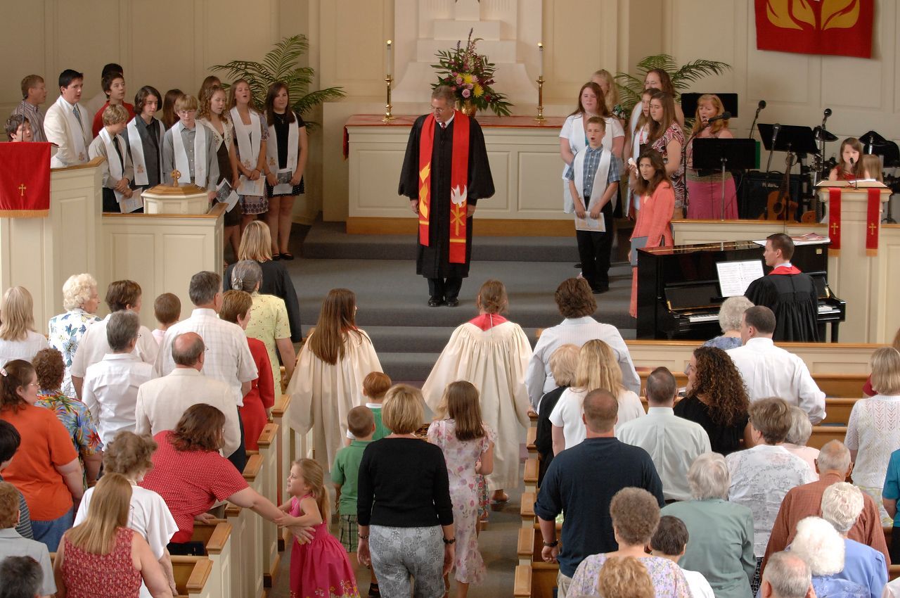 Church service with people in pews, a pastor, and choir, with piano and red cross symbol.