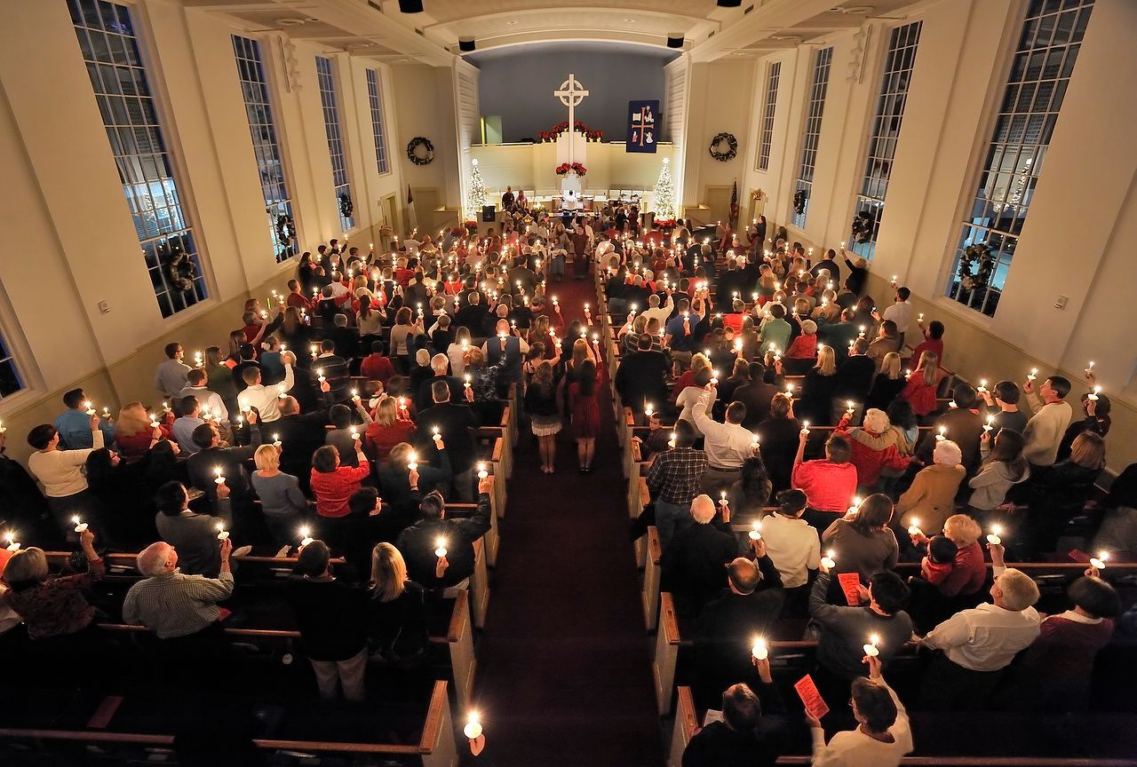 People in a church holding lit candles. Central aisle with figures walking. Many lit candles fill the space.