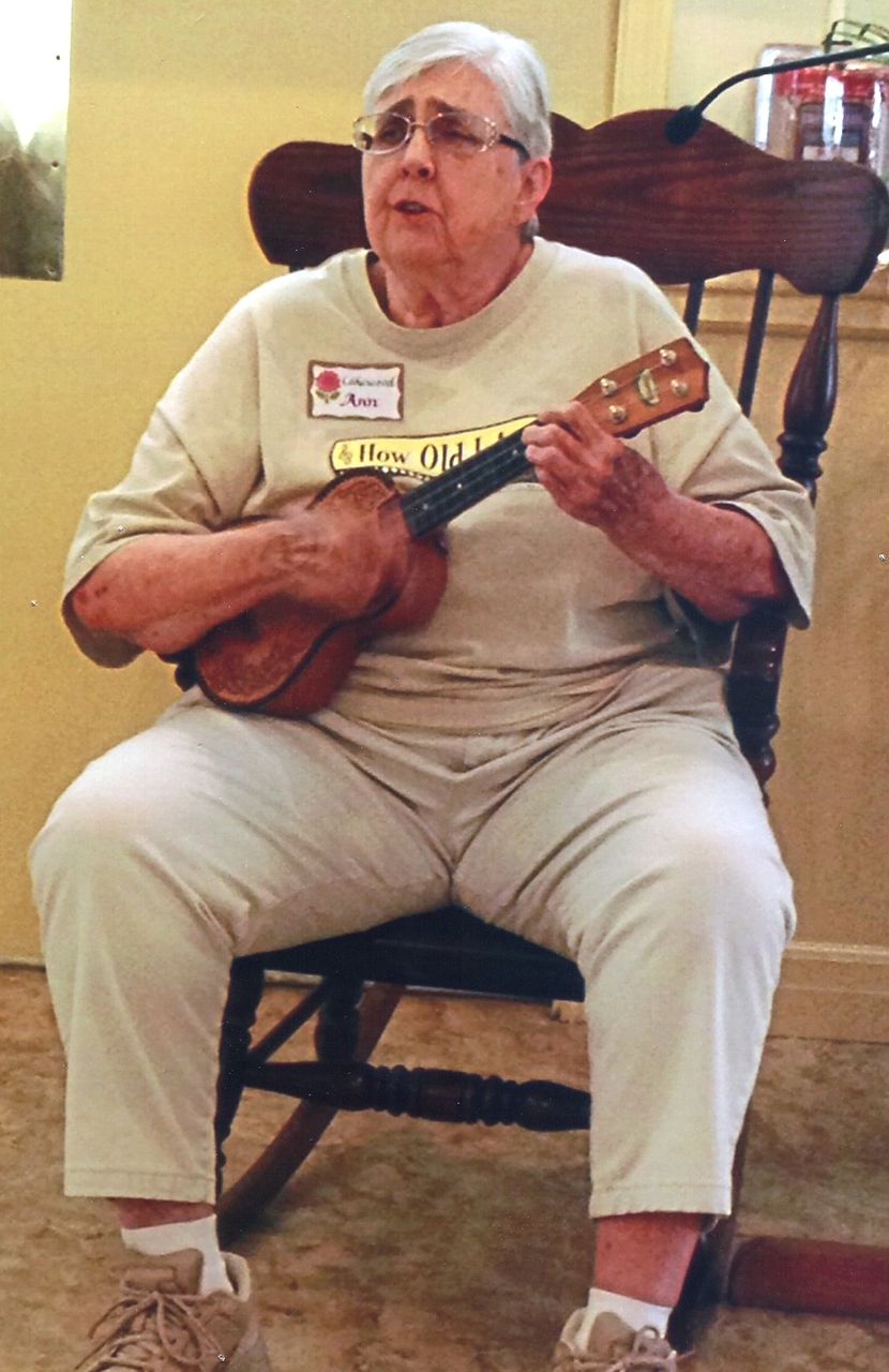 Woman seated, playing a ukulele, indoors, wearing light-colored clothing.