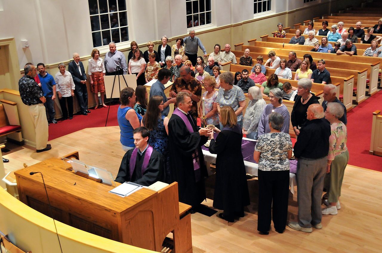 People gathering for a church service. A priest in purple robes is interacting with people gathered around an altar.
