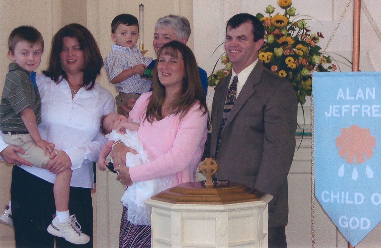 Family at a baptism ceremony: woman holding baby, people around a wooden font, banner on the right.