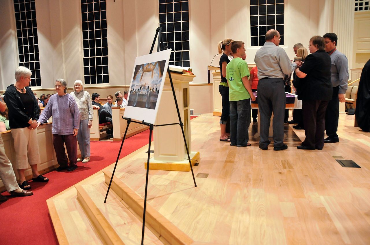 People gather in a church, looking at a framed photo on an easel. Wooden floor and steps, white walls and a red carpet.